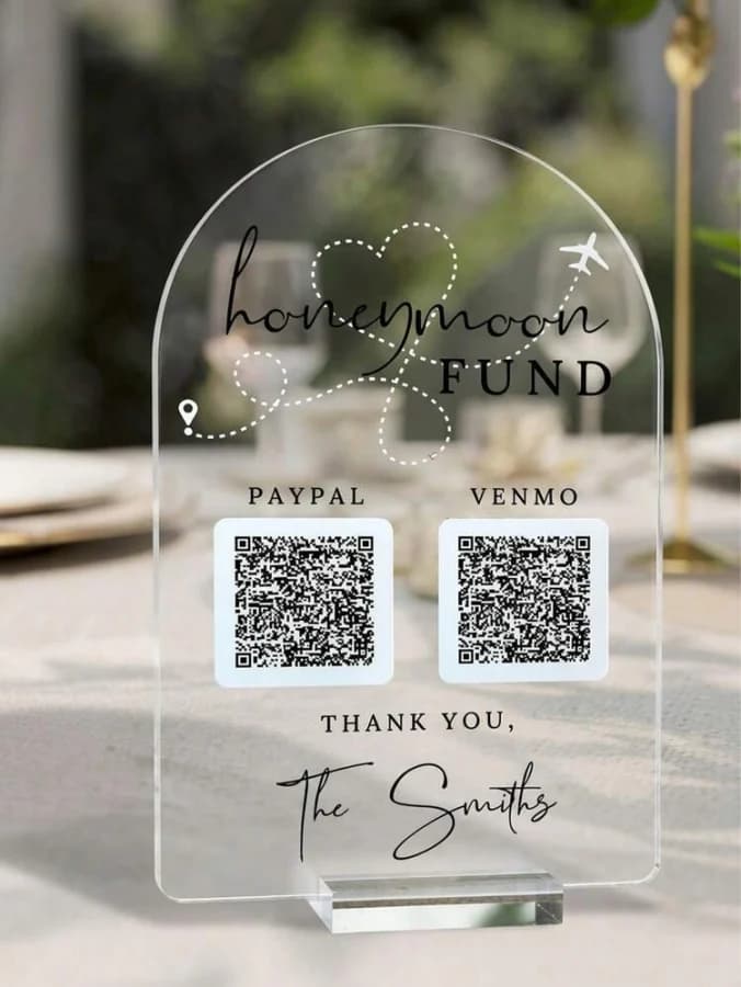 A clear acrylic arch sign on a laid table reading Honeymoon Fund, with two scannable codes labelled PayPal and Venmo